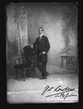 Foto de estudio de un varón que se encuentra parado y apoyado a un sillón. Lleva puesto una camisa con cuello almidonado, corbata y traje frac. Sostiene en mano unos guantes y un bastón.
