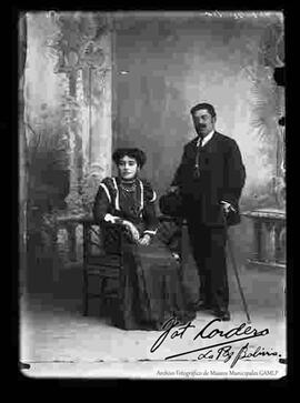 Foto de estudio de una pareja. Una dama que se encuentra sentada en una silla y que lleva puesto un vestido de seda largo y de color oscuro. El varón se encuentra parado y presenta lentes y un traje formal con chaleco. Sostiene en mano un sombrero bombín y un bastón.