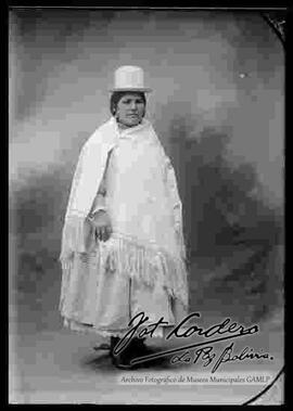 Foto de estudio de una chola que se encuentra parada. Lleva puesto un sombrero blanco de copa alta, una manta, pollera con centro sobresalido y botines con tacón