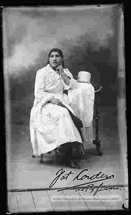 Foto de estudio de una chola que se encuentra   sentada, con el brazo en el mentón, apoyada en una mesita donde estan puestos, su sombrero de copa alta y color blanco asi como su manta. Lleva puesto una blusa de manga larga con encajes, pollera y botines con tacón.