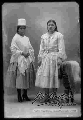 Foto estudio de dos cholas que se encuentran paradas. Una lleva puesto un sombrero de copa alta, manta, pollera y botines con tacón y la otra lo mismo con la diferencia de la ausencia del sombrero