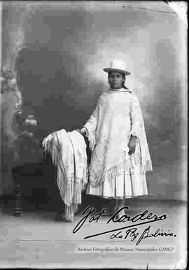 Foto de estudio de una chola que se encuentra parada y apoyada a un  sillón donde esta una manta de seda. Lleva puesto un sombrero blanco de copa alta, manta de lana tejida,  pollera a rayas y botines con tacón