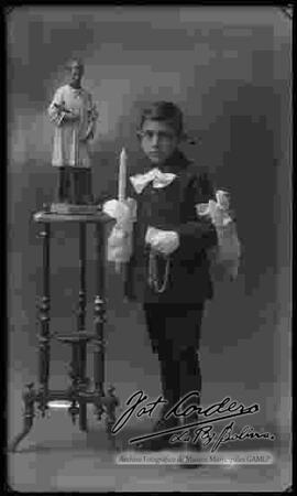 Foto de estudio de un niño del colegio La Salle, el día de su primera comunión, parado a lado de la imagen de un santo