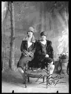 Foto de estudio de dos señoritas que se encuentran sentadas en una silla y llevan puesto; Un tipo de sombrero, abrigos de piel y faldas hasta la rodilla, con zapatillas con tacón.