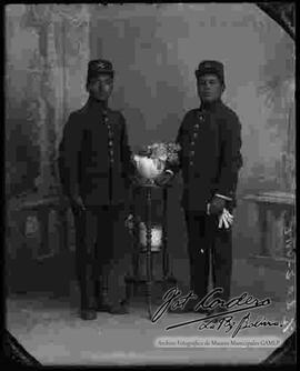 Foto de estudio de dos soldados, apoyados a una mesita con flores