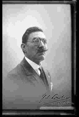 Foto de estudio de medio cuerpo de un varón de lentes y bigote, que viste de traje.