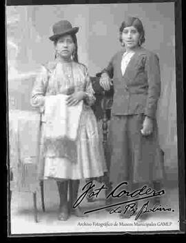 Foto de estudio de una chola que se encuentra parada a lado de una dama y una mesita donde esta una manta. Lleva puesto un sombrero de color oscuro, blusa de seda y de media manga, pollera y botines con tacón. Sostiene en mano una manta bordada.