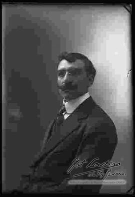 Foto de estudio de un varón de lentes y bigote que viste de traje.
