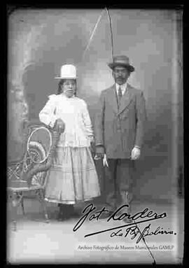 Foto de estudio de una chola junto a su pareja.  Se encuentra apoyada a una silla de mimbre, con sombrero blanco de copa alta, blusa larga y ancha, pollera con enaguas sobresalidas y botines con tacón. El varon es un hombre criollo o cholo de bigotes con sombrero y traje