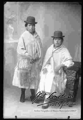 Foto de estudio de dos cholas, una se encuentra sentada y la otra parada. Ambas llevan puesto: Sombreros borsalinos, manta y pollera. Una con botines de tacón  y la otra con zapatillas planas.