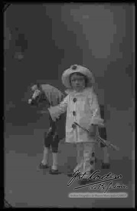 Foto de estudio de un niño disfrazado de arlequín, parado junto a un caballito de juguete.