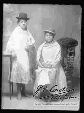 Foto de estudio de dos cholas. Una se encuentra parada, apoyada a una mesita y la otra, sentada en un sillón. Ambas lucen sombreros borsalinos de color oscuro, mantas y polleras. Una con botines de tacón y la otra con zapatillas