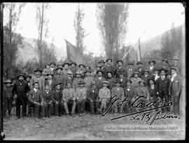 Grupo de varones y hacendados de una provincia, reunidos en el patio de una casa particular, sosteniendo dos banderas en el fondo