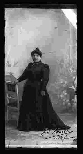 Foto de estudio de una dama que se encuentra parada y apoyada a una silla. Lleva puesto un vestido largo de color oscuro y un collar con una cruz.