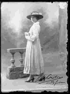 Foto de estudio de una dama parada junto a un barandal, lleva puesto: Un sombrero, un vestido largo de color claro y botines con tacón.