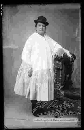 Foto de estudio de una chola que se encuentra parada y apoyada a un sillón. Luce un sombrero borsalino color negro, manta sujeta por un topo pequeño, pollera y botines con tacón