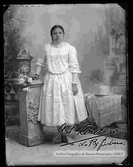 Foto estudio de una chola de cuerpo entero y apoyada a un mueble. Lleva puesto una blusa de manga larga, pollera y botines de tacón, presenta a si mismo al lado en una mesa el sombrero de copa alta y una manta