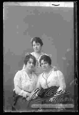 Foto de estudio de tres damas. Dos se encuentran sentadas y una parada. Visten blusas blancas de manga larga y faldas largas de color oscuro