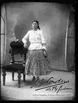 Foto de estudio de una chola que se encuentra parada y apoyada a un sillón, sin sombrero. Lleva puesto una blusa cerrada de manga larga, pollera floreada, botines, un collar con libra esterlina y aretes.