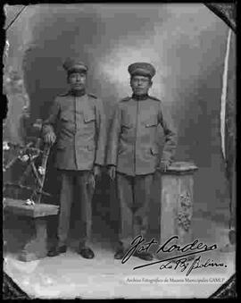 Foto de estudio de dos soldados, parados a lado de una pequeña columna y una banca