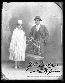 Foto de estudio de una chola junto a un caballero. La mujer luce un sombrero de copa alta, manta pollera y botines de tacón. El varon lleva un sombrero y traje con chaqueta