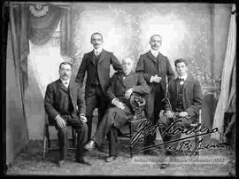 Foto de estudio de un grupo de cinco varones vestidos de traje. Tres sentados en sillas y dos parados por detrás