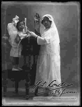 Foto de estudio de una niña, el día de su primera comunión, junto a una niña pequeña vestida de ángel, parado encima de un sillón