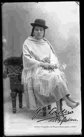 Foto de estudio de una chola que se encuentra sentada en un sillón y agarrando flores. Lleva puesto un sombrero borsalino de color negro, manta, pollera y botines con tacón