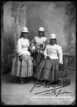 Foto de estudio de tres cholas. Dos se encuentran paradas y una sentada en un mueble. Llevan puesto sombreros de copa alta y de color blanco, blusas cerradas de manga larga, pollera y botines con tacón