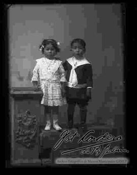 Foto de estudio de dos niños. Una niña y un  niño, parados encima de una pequeña pared tallada de madera junto a una columna