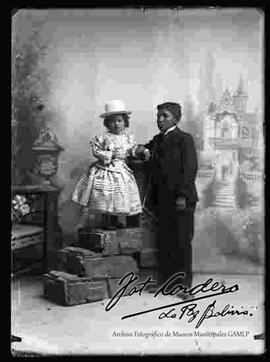 Foto de estudio de una niña cholita, parada encima de una pequeña pared junto a su hermano. Lleva puesto un sombrero blanco de copa alta, una blusa de manga larga con encajes, pollera y botines