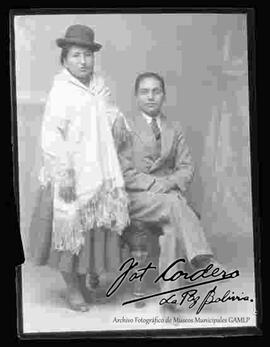 Foto de estudio de una pareja. Una chola que se encuentra parada junto a su esposo que esta sentado en un sillón. Lleva puesto un sombrero borsalino de color oscuro, manta bordada, pollera y zapatillas planas