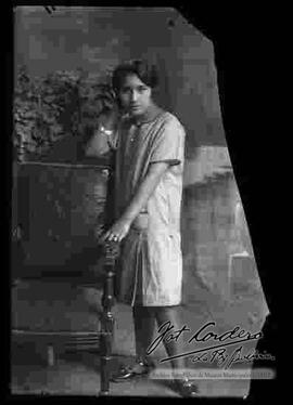 Foto de estudio de una joven señorita que se encuentra parada y apoyada a una silla. Lleva puesto un vestido de manga corta y zapatos planos.