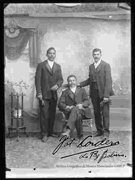 Foto de estudio de tres varones vestidos de traje. Dos se encuentran parados y uno sentado en medio, en una silla de mimbre