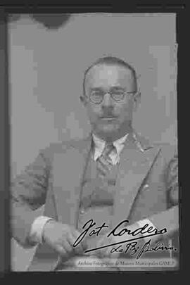 Foto de estudio de medio cuerpo de un varón de lentes, que se encuentra sentado y que viste de traje