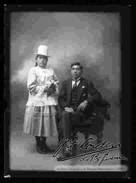 Foto de estudio de una pareja. Una chola que se encuentra parada junto a su esposo. Ella lleva puesto un sombrero blanco de copa alta, blusa de manga larga con encajes, pollera y botines con tacón.