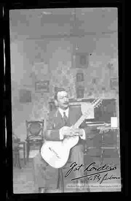 Musico sentado en una silla, sosteniendo una guitarra, en el interior de la sala de una casa.
