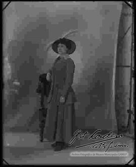 Foto de estudio de una dama apoyada a un sillón, lleva puesto un traje de dos piezas combinado con un modelo de sombrero.