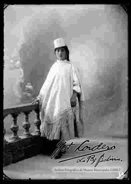Foto de estudio de una chola que se encuentra parada y apoyada a una baranda. Lleva puesto un sombrero blanco de copa alta, manta bordada, pollera a rayas y botines con tacón de color negro.