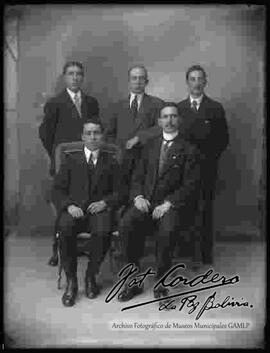 Foto de estudio de cinco varones que visten de traje. Dos se encuentran sentados  y tres parados.
