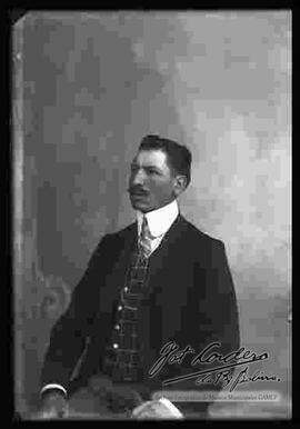 Foto de estudio de un varón que se encuentra sentado. Lleva puesto un traje formal con chaleco a cuadros, camisa blanca y corbata.