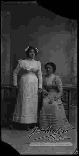 Foto de estudio de dos damas. Una se encuentra parada y la otra sentada en una silla