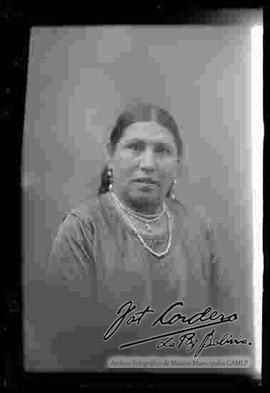 Foto de estudio de medio cuerpo, de una chola sin sombrero. Lleva puesto aretes, un collar de perlas y una blusa cerrada bordada