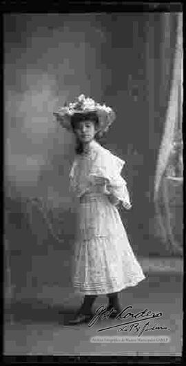 Foto de estudio de una dama  que lleva puesto, un sombrero con encajes y un vestido largo.