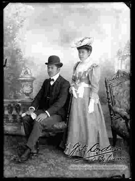 Foto de estudio de una pareja. Un varón que se encuentra sentado en una silla y que lleva puesto: Un sombrero bombín, un traje con chaleco y guantes. La dama se encuentra parada y lleva puesto un sombrero con adornos, un vestido largo y guantes en las manos.