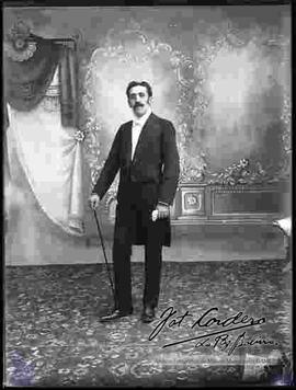 Foto de estudio de un varón que lleva puesto: Una camisa blanca, corbata de gato, traje de fiesta o smoking. Sostiene en mano unos guantes y un bastón.