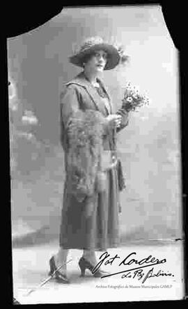 Foto de estudio de una dama lleva puesto: Un sombrero con plumas, vestido largo  sujeto por una cinta en la cadera y sosteniendo  en mano una estola de zorro.