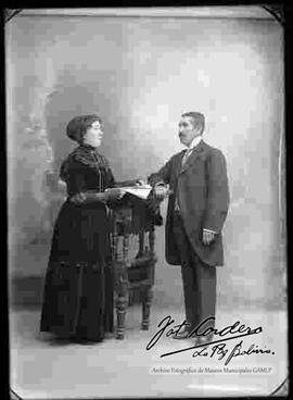 Foto de estudio de una pareja apoyados a un sillón. La  dama sostiene una revista y lleva puesto un vestido largo de cuello cerrado y de color oscuro. El varón lleva puesto un traje formal.