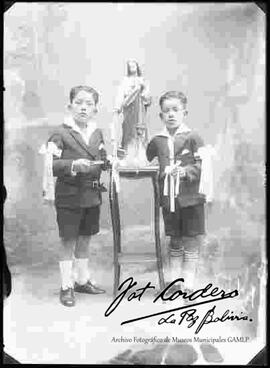 Foto de estudio de dos niños junto a una imagen religiosa, en el día de su primera comunión