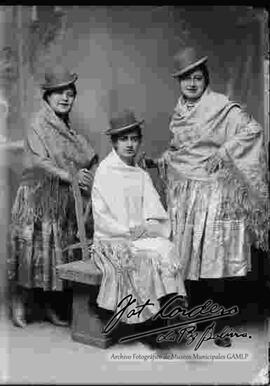 Foto de estudio de tres jóvenes vestidas de cholas. Dos se encuentran paradas y una sentada en medio en una banca. Llevan puesto sombreros borsalinos de color oscuro, mantas bordadas, polleras y botines con tacón.